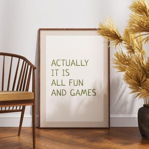 May include: A framed print with the text "ACTUALLY IT IS ALL FUN AND GAMES" in green. The frame is brown, and the background is a light beige. The print is leaning against a white wall, next to a wooden chair and a vase of dried plants.