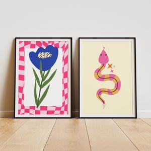 May include: Two framed art prints. The left print features a blue flower with green leaves against a white background, framed by a pink and white checkered border. The right print shows a pink and yellow snake with star accents on a cream background.