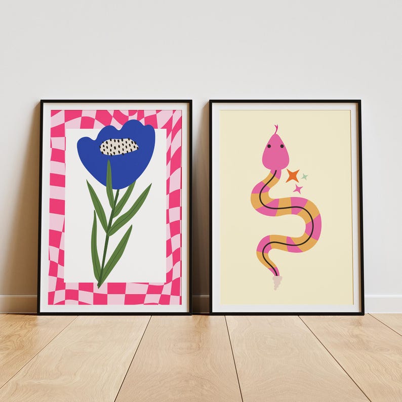 May include: Two framed art prints. The left print features a blue flower with green leaves against a white background, framed by a pink and white checkered border. The right print shows a pink and yellow snake with star accents on a cream background.