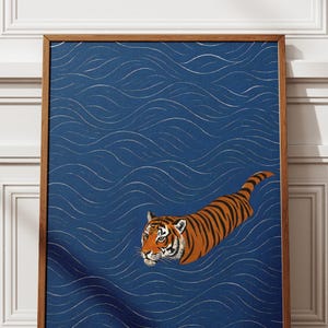 Puede incluir: Una impresi&oacute;n art&iacute;stica enmarcada que representa un tigre nadando en agua azul con patrones de olas blancas. El tigre tiene rayas naranjas y negras y mira hacia la izquierda. El marco es de madera marr&oacute;n.
