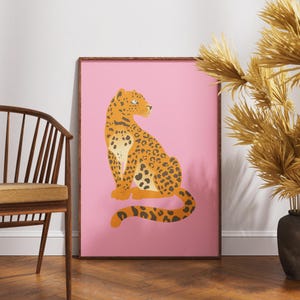 May include: A framed art print featuring a stylized leopard illustration. The leopard is orange with black spots, set against a pink background. The frame is brown. The artwork is displayed near a wooden chair and decorative dried plants.