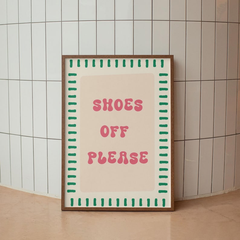 May include: A rectangular wall art print in a brown wooden frame featuring a light beige background with a green dashed border. The text "SHOES OFF PLEASE" is printed in the center in a bold, pink, wavy-style font, creating a clear visual message.