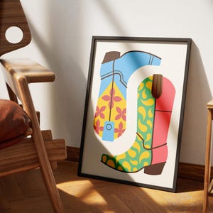 May include: A framed art print featuring a stylized illustration of two cowboy boots. One boot is blue with yellow and pink floral accents, the other is red and green with a yellow paisley pattern. The boots are set against a cream background.