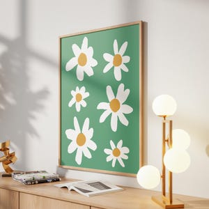 May include: A framed art print featuring a green background with white and yellow daisy illustrations. The artwork is displayed on a wooden surface, alongside a modern lamp with spherical lights and decorative objects.