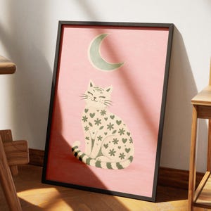 Puede incluir: Una impresión artística enmarcada con una ilustración caprichosa de un gato. El gato está adornado con patrones de corazones y flores, sobre un fondo rosa suave. Una luna creciente está encima del gato. La obra de arte está en un marco negro.
