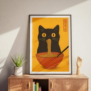 May include: A framed art print featuring a black cat eating noodles from a red bowl. The cat has yellow eyes and is depicted against a yellow background. The print includes Japanese text in a red box. A black chopstick is in the bowl.