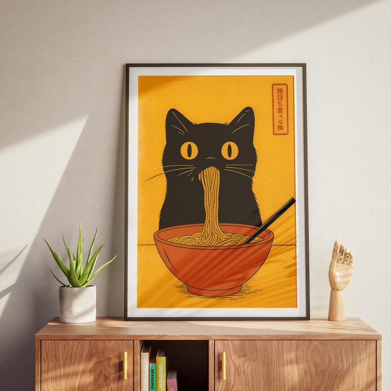May include: A framed art print featuring a black cat eating noodles from a red bowl. The cat has yellow eyes and is depicted against a yellow background. The print includes Japanese text in a red box. A black chopstick is in the bowl.