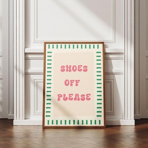May include: A rectangular framed wall art piece featuring a cream-colored background with the pink text "SHOES OFF PLEASE" in a rounded font. The border consists of a repeating pattern of short, vertical green dashes on a white background.