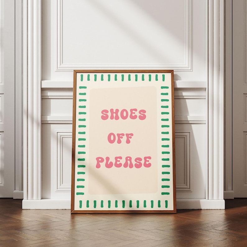 May include: A rectangular framed wall art piece featuring a cream-colored background with the pink text "SHOES OFF PLEASE" in a rounded font. The border consists of a repeating pattern of short, vertical green dashes on a white background.