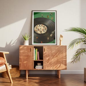 Puede incluir: Una impresi&oacute;n art&iacute;stica enmarcada representa un disco de vinilo con peque&ntilde;as figuras bailando sobre &eacute;l, sobre un fondo verde. La obra de arte se exhibe sobre un mueble de madera con tiradores dorados, una peque&ntilde;a planta y libros.