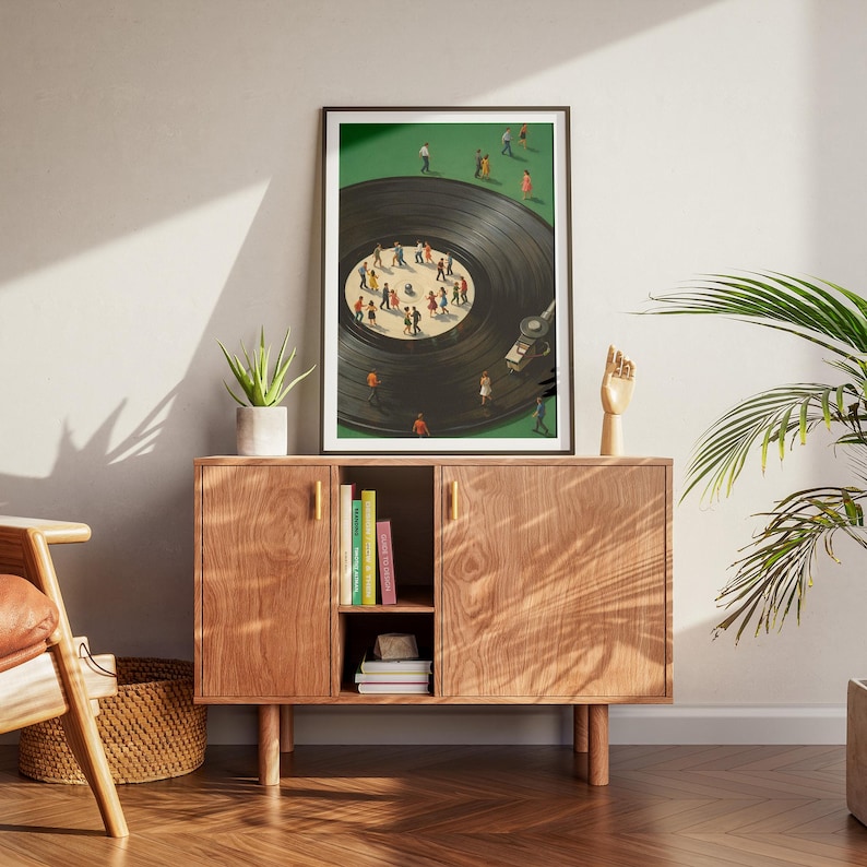 Puede incluir: Una impresi&oacute;n art&iacute;stica enmarcada representa un disco de vinilo con peque&ntilde;as figuras bailando sobre &eacute;l, sobre un fondo verde. La obra de arte se exhibe sobre un mueble de madera con tiradores dorados, una peque&ntilde;a planta y libros.