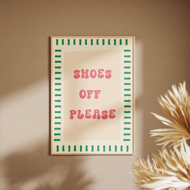 May include: A rectangular wall print in a light wooden frame featuring a cream-colored background with a green dashed border. The text "SHOES OFF PLEASE" is printed in the center in a bold, pink, rounded font, conveying a clear message.