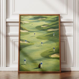 Puede incluir: Pintura enmarcada de un campo de golf con golfistas con camisas azules y pantalones blancos. El campo verde tiene trampas de arena y terreno ondulado. La obra de arte está en un marco de madera, apoyada en una pared blanca y un suelo de madera.