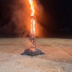 May include: A metal fire pit with flames and smoke rising into the night sky. The fire pit is square at the base and has a decorative design on the sides. The ground is covered in dirt and small stones.