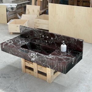 K&ouml;nnte beinhalten: Ein Badezimmerwaschtisch aus dunkelrotem Marmor mit Doppelwaschbecken und einem wei&szlig;en Seifenspender. Der Waschtisch wird von einer Holzpalette getragen. Der Marmor hat wei&szlig;e Adern und eine strukturierte, geriffelte Kante.