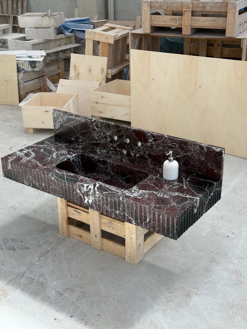 K&ouml;nnte beinhalten: Ein Badezimmerwaschtisch aus dunkelrotem Marmor mit Doppelwaschbecken und einem wei&szlig;en Seifenspender. Der Waschtisch wird von einer Holzpalette getragen. Der Marmor hat wei&szlig;e Adern und eine strukturierte, geriffelte Kante.