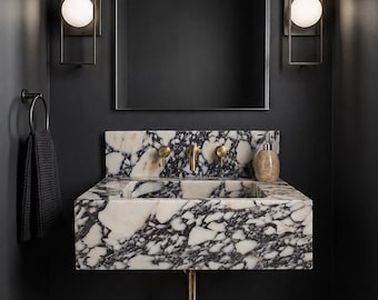 May include: A wall-mounted rectangular sink crafted from white and black veined marble, featuring brass faucets and handles. Above the sink is a rectangular mirror flanked by two black metal wall sconces with spherical white glass globes.