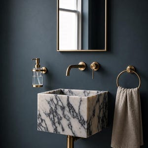 May include: A wall-mounted square marble sink with black veining, paired with a brass faucet, handle, soap dispenser, and circular towel ring holding a beige towel. A rectangular mirror with a thin brass frame is mounted above the sink.