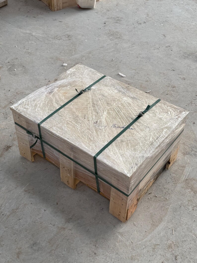 K&ouml;nnte beinhalten: Eine rechteckige Holzpalette mit einem verpackten Paket. Das Paket ist mit gr&uuml;nen Gurten und klarer Plastikfolie gesichert. Die Palette besteht aus hellem Holz und steht auf einem Betonboden.