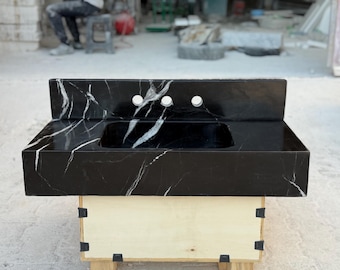 May include: A black marble bathroom vanity sink featuring prominent white veining throughout the stone surface. The rectangular basin is integrated into the countertop, which includes a matching backsplash with three pre-drilled faucet holes.