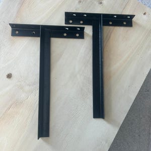 May include: Two black metal shelf brackets in an L-shape, with multiple screw holes for mounting. The brackets are positioned on a light-colored wooden surface, showcasing their design and potential use for shelving.