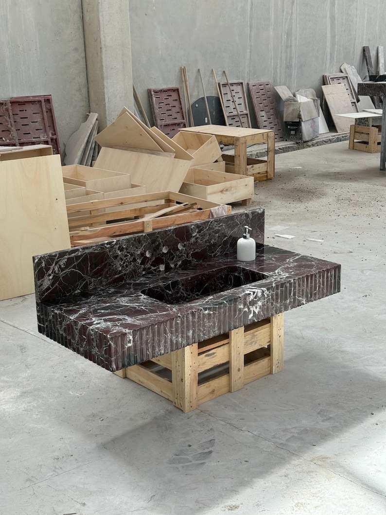K&ouml;nnte beinhalten: Eine dunkelrote Marmor-Arbeitsplatte mit eingebautem Waschbecken und einem wei&szlig;en Seifenspender. Die Arbeitsplatte wird von einer Holzpalettenbasis getragen. Der Marmor hat wei&szlig;e Adern und eine strukturierte Oberfl&auml;che.