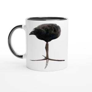 May include: White ceramic coffee mug with a black rim and handle. The mug features a whimsical illustration of a black bird with a long leg and three-toed foot. The bird's body is round and fluffy.