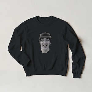 May include: Black sweatshirt featuring a black and white image of a person wearing a hat and smiling widely. The image is centered on the front of the long-sleeved crew neck sweatshirt.