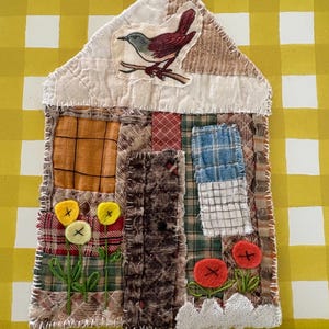 Peut inclure: Une décoration murale en tissu en forme de maison composée de divers carrés de patchwork dans des tons marron, bleu et orange. Elle présente un appliqué d'oiseau, des fleurs brodées jaunes et rouges, et des coutures blanches. 12,7 cm de large.