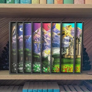 May include: A collection of fantasy books in a black case, presented on a wooden shelf. The book covers showcase colourful illustrations of dragons, castles, and landscapes with a deer. The books are arranged in a row.
