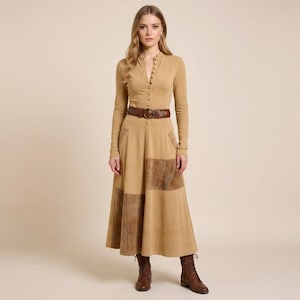 May include: A tan-colored long-sleeved dress with a button-up top and a long skirt. The skirt features plaid patches and is cinched at the waist with a brown belt. The model is wearing brown boots.