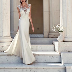 Puede incluir: Un vestido de novia blanco con un corpiño ajustado y una falda fluida. El corpiño presenta delicados detalles de encaje en los hombros y el escote. El vestido tiene una sutil cola y se usa al aire libre en escaleras de mármol.