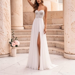 May include: A white, strapless wedding dress with a high leg slit. The bodice features lace detailing, and the skirt falls to the floor. The setting is outdoors, with stone columns and steps.