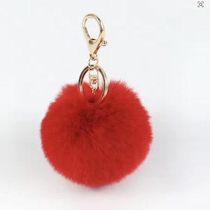 May include: A bright red fluffy pom-pom keyring with a gold-coloured clasp and ring. The keyring is round and appears soft and plush. The gold hardware provides a contrasting accent to the bright red pom-pom.