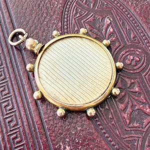 May include: A round gold-toned pendant featuring a central disc with a fine diagonal engraved texture. The circular frame is accented with eight small spherical beads along the outer edge and includes a small jump ring at the top for attachment.