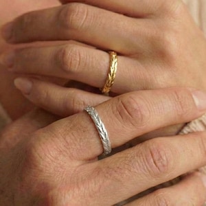 May include: Two rings with a wheat pattern are shown. One ring is gold, and the other is silver. The rings are on fingers, suggesting a theme of commitment or adornment. The background is neutral, emphasising the jewellery.