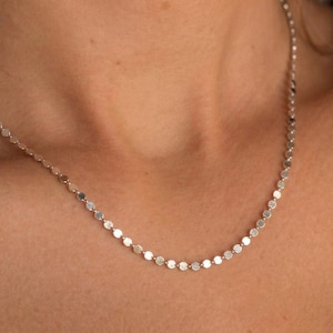 Può includere: Una delicata collana d'argento con piccoli ciondoli rotondi e piatti. La collana è mostrata su uno sfondo colorato, evidenziando la sottile lucentezza e il design minimalista del gioiello. La collana è un accessorio semplice ma elegante.