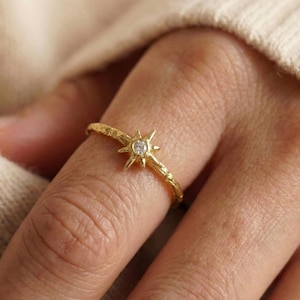 Peut inclure: Une bague dorée avec un motif d'éclat d'étoile et une petite pierre précieuse transparente. La bague est portée au doigt et possède un anneau texturé. Il s'agit d'un bijou.