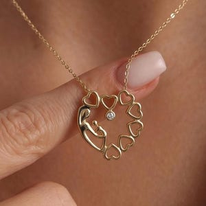 Peut inclure: Un collier doré avec un pendentif en forme de cœur. Le pendentif représente une mère et son enfant, entourés de petits cœurs, avec une petite pierre précieuse transparente au centre. Le collier est suspendu à une fine chaîne.