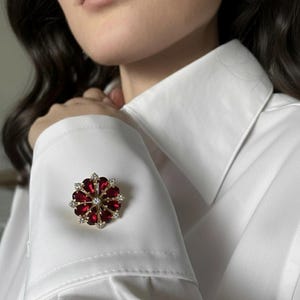 May include: A gold-toned cufflink with a floral design. The cufflink features a central clear stone surrounded by red, heart-shaped stones and smaller clear stones. The cufflink is set against a white dress shirt.