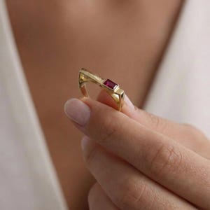May include: A gold ring featuring a rectangular, deep red gemstone. The ring has a distinctive, angular design, with a band that curves upwards. The gemstone is set in a bezel, and the ring is held in a hand.