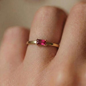 Op de afbeelding: Een gouden ring met een rechthoekige, helderrode edelsteen. De ring heeft een slanke, gepolijste band, met de edelsteen gezet in een eenvoudig, elegant ontwerp. De ring wordt op een vinger getoond, waarbij de delicate details worden benadrukt.