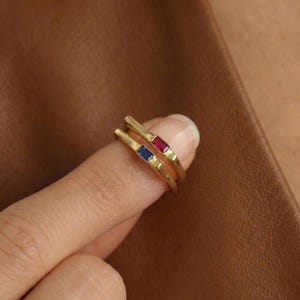 May include: Two gold rings, one with a red gemstone and the other with a blue gemstone. The rings are thin bands with rectangular stones. The rings are displayed on a finger against a brown background.