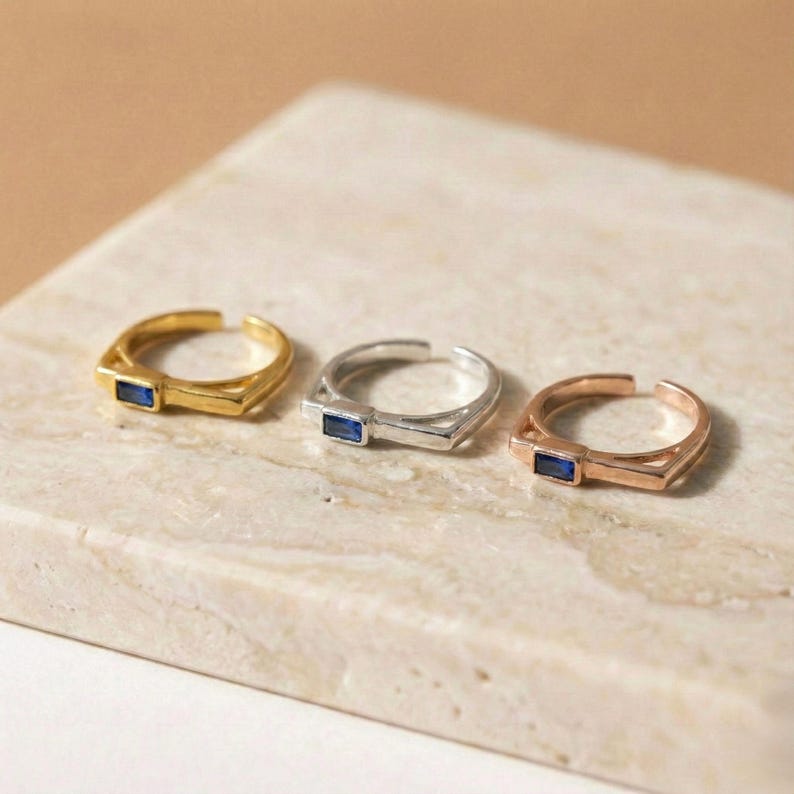 May include: Three adjustable rings in gold, silver, and rose gold, each with a rectangular blue gemstone. The rings are presented on a light-coloured, textured surface, highlighting their minimalist design and metallic finishes.