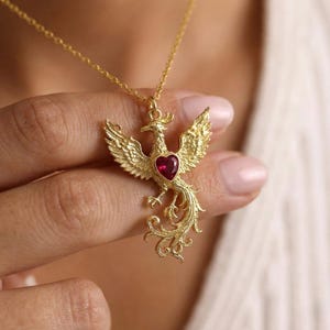 May include: A gold phoenix pendant necklace with a red heart-shaped gemstone. The phoenix has outstretched wings and detailed feathering. The necklace is suspended from a delicate gold chain.