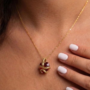 Könnte beinhalten: Eine goldfarbene Halskette mit einem Anhänger, der einen roten Edelstein zeigt, der von einer goldfarbenen Schlange umschlungen ist. Die Halskette hat eine feine goldfarbene Kette. Der Anhänger hat einen Durchmesser von ca. 2 cm.