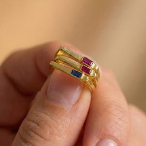 May include: Three gold rings stacked together, each with a rectangular gemstone. The top two rings have red gemstones, and the bottom ring features a blue gemstone. The rings are held in a hand.