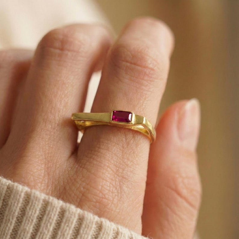 May include: A gold ring with a rectangular, deep red gemstone. The ring has a modern, minimalist design with a smooth, polished finish. The gemstone is set in a bezel setting, and the band has a unique, sculpted shape.