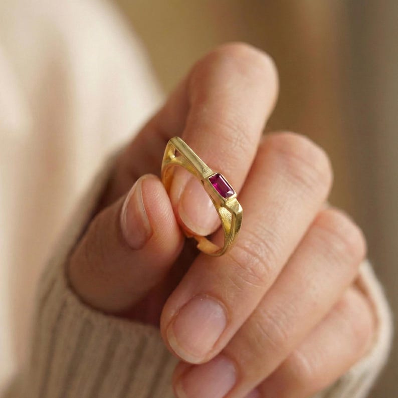 A minimalist 14k gold plated ring featuring a rectangular red ruby in a unique geometric bar setting held by a hand.