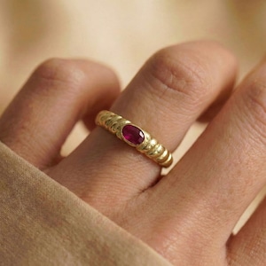 May include: A gold ring with a textured band and a central oval ruby gemstone. The ring is a warm gold color and has a ribbed design around the band.
